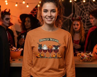 Può includere: Una maglietta arancione a maniche lunghe con una grafica di quattro calici da vino pieni di liquido rosso. All'interno dei bicchieri ci sono un gatto nero, un fantasma, una zucca e un pipistrello. Il testo sopra recita Drink Up Witches.