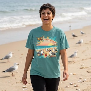 May include: A teal short-sleeve t-shirt featuring a graphic of seagulls on a beach with sunglasses, a floatie, and chips. Above the scene, a small aeroplane flies near a sun with the text, "IT'S A GOOD DAY FOR A GOOD DAY!"