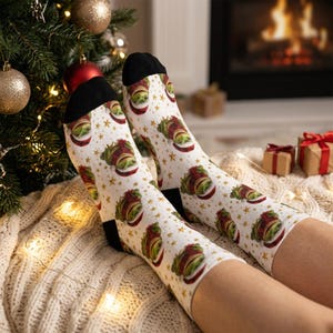 May include: White crew socks featuring a repeating pattern of green creatures wearing red and white Santa hats, interspersed with small gold stars. The socks have solid black cuffs, heels, and toes, providing a festive look for the holiday season.