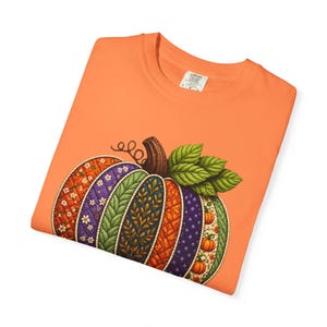 May include: An orange t-shirt featuring a large graphic of a quilted pumpkin. The pumpkin is composed of vertical segments with various patterns including floral, polka dot, and leaf designs in shades of purple, green, and orange with green leaves.