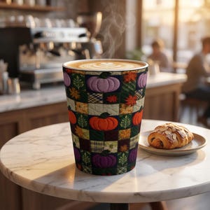 Peut inclure: Un grand gobelet orné d'un motif patchwork matelassé représentant des citrouilles orange et violettes, des feuilles vertes et des carrés à carreaux. Il contient un latte fumant avec de l'art latte, posé près d'une pâtisserie aux amandes.
