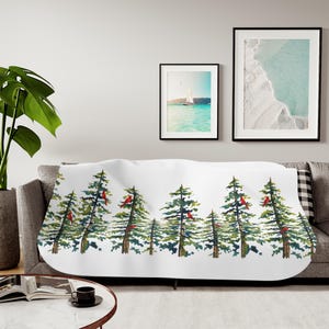 May include: A white throw blanket featuring a watercolor-style print of green pine trees and small red cardinal birds. The pattern is arranged in a horizontal row across the center of the fabric, showcasing detailed brushstrokes and natural colors.