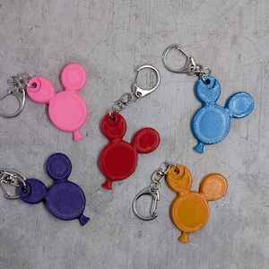 以下が含まれることがあります： ミッキーマウスの風船の形をした、カラフルなキーホルダーのコレクション。キーホルダーはピンク、赤、青、紫、黄色です。各キーホルダーには、鍵やバッグに取り付けるためのシルバーの留め金が付いています。キーホルダーはテクスチャード加工された素材で作られています。
