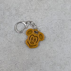 以下が含まれることがあります： シルバーの留め金が付いた、タン色のミッキーマウスのキーホルダー。キーホルダーは、ミッキーマウスの顔の輪郭が詳細に描かれており、耳、目、鼻、口が含まれています。キーホルダーは丈夫な素材で作られています。