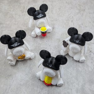 May include: Four white figurines with black mouse ears and hats. Each figurine has a different accessory, including a coffee cup, a star, and a brown shape. The figurines are on a grey surface.