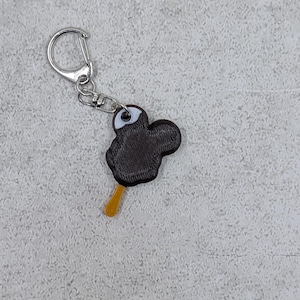 以下が含まれることがあります： 銀色の留め金が付いた、黒いアイスキャンディーのキーホルダー。アイスクリームは白と茶色のデザインで、黄色の棒が付いています。楽しいアクセサリーです。