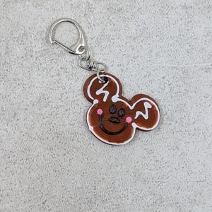 以下が含まれることがあります： 茶色のジンジャーブレッド風キーホルダー。漫画のネズミの形をしています。キーホルダーはシルバーの留め金と、笑顔の顔、ピンクの頬、白いアイシングのディテールが付いた小さくて丸いチャームが付いています。チャームの幅は約5cmです。