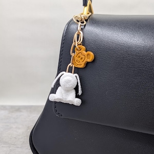 May include: A black leather handbag featuring a gold-toned metal chain charm. The charm includes a small, tan-colored, molded character head with rounded ears and a white, plush, soft-textured rabbit figure with long ears attached to the chain.