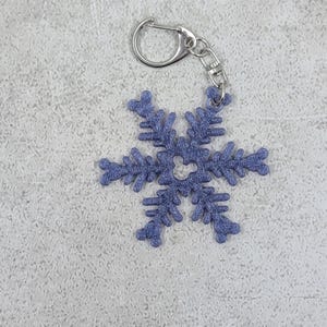 以下が含まれることがあります： シルバーのキーリングと留め金が付いた、青い雪の結晶のキーホルダー。 雪の結晶は、複雑な模様と質感のある表面が特徴で、冬をテーマにしたアクセサリーやギフトに最適です。