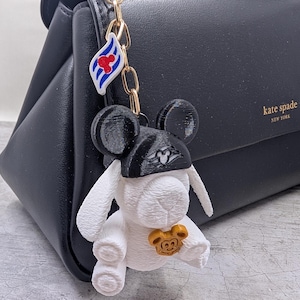 Op de afbeelding: Een witte konijn-sleutelhanger met een zwarte pet met een gevleugeld embleem, een goudkleurige Mickey Mouse-hoofdbedeltje en een kleine witte hanger met blauwe strepen en een rode Mickey Mouse-kop aan een goudkleurige metalen ketting.