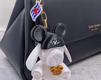 Puede incluir: Un llavero de conejo blanco con una gorra negra que presenta un emblema estilo Mickey Mouse, un pequeño dije de cabeza de Mickey Mouse color dorado y un dije de bandera a rayas blancas y azules con una cabeza de Mickey Mouse roja, en una cadena dorada.