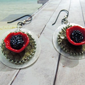 Peut inclure: Une paire de boucles d'oreilles avec une fleur de coquelicot rouge au centre noir, posée sur une base circulaire argentée et dorée avec un bord perlé.