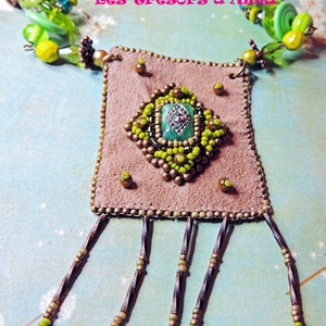 Könnte beinhalten: Ein brauner Wildlederanhänger mit einem grünen und goldenen Perlenmuster und einem grünen Edelstein in der Mitte. Der Anhänger ist an einer braunen Schnur mit grünen und goldenen Perlen befestigt. Der Anhänger hat Fransen an der Unterseite.