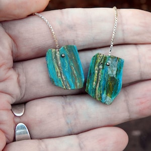 Op de afbeelding: Een paar oorhangers met turquoise en goudkleurige stenen accenten. De oorbellen zijn gemaakt met zilveren kettingen en hebben een unieke, natuurlijke uitstraling.