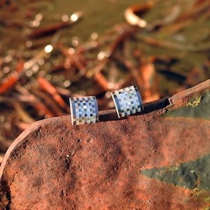 Peut inclure: Deux boucles d'oreilles carrées argentées avec un motif à carreaux, reposant sur une surface métallique rouillée.