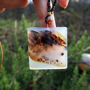 Puede incluir: Collar con colgante cuadrado de piedra blanca translúcida con patrones marrones y negros. El colgante cuelga de un collar de cuentas negras y un cordón de cuero marrón. Fondo verde borroso.