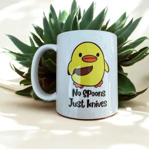 May include: White ceramic mug with a cartoon yellow chick holding a knife. The mug has the text "No Spoons Just Knives" in black. The mug is set against a backdrop of green succulent leaves.