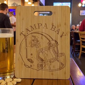 May include: A wooden cutting board with the words "TAMPA BAY" and a football helmet design etched into the surface. The helmet features a skull and crossbones logo. The board has a handle and is sitting on a wooden table.