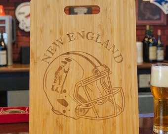 May include: A rectangular light brown bamboo chopping board featuring a laser-engraved design of a football helmet with a colonial figure and the text NEW ENGLAND arched above. The board includes a centred rectangular handle cutout at the top.