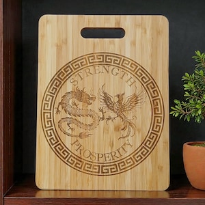 May include: A rectangular bamboo cutting board with a handle. The board features an engraved design of a dragon and phoenix within a circular border, with the words "STRENGTH" and "PROSPERITY".