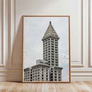 May include: A framed print of a tall, light-colored building with a distinctive triangular top. The building has many windows and is set against a cloudy sky. The frame is a light brown color, and the print is displayed on a light-colored wall.