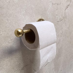 May include: A gold-colored metal toilet paper holder with a roll of white toilet paper. The holder is mounted on a light gray marble-patterned wall. The holder has a round ball on the end.