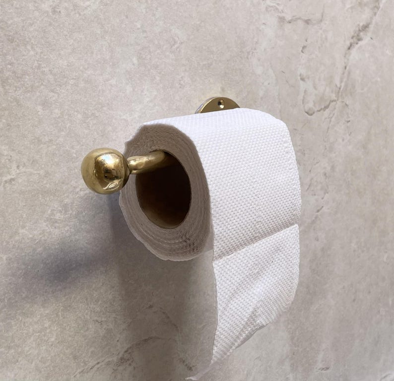 May include: A gold-colored metal toilet paper holder with a roll of white toilet paper. The holder is mounted on a light gray marble-patterned wall. The holder has a round ball on the end.