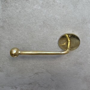 May include: A gold-colored, wall-mounted toilet paper holder. The holder features a rounded ball on one end, a straight bar, and an oval-shaped mounting plate. The fixture is made of a shiny, metallic material.