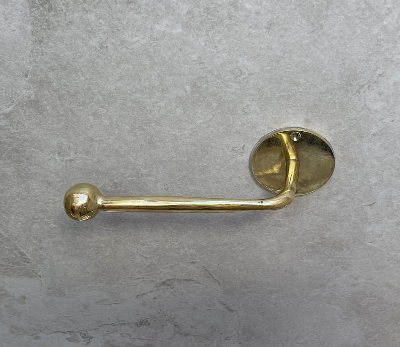 May include: A gold-colored, wall-mounted toilet paper holder. The holder features a rounded ball on one end, a straight bar, and an oval-shaped mounting plate. The fixture is made of a shiny, metallic material.