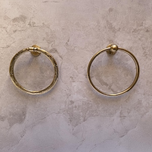 May include: Two brass towel rings mounted on a light gray marble-patterned wall. One ring has a textured surface, while the other is smooth with a small ball detail at the top. Both are circular and designed for bathroom use.