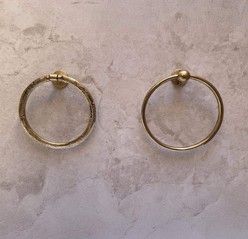 May include: Two brass towel rings mounted on a light gray marble-patterned wall. One ring has a textured surface, while the other is smooth with a small ball detail at the top. Both are circular and designed for bathroom use.