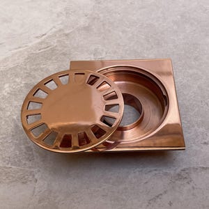 Könnte beinhalten: Ein kupferfarbener quadratischer Abdeckrost mit einem kreisförmigen, geschlitzten Deckel. Der Deckel ist teilweise entfernt und gibt das Innere des Ablaufs frei. Der Ablauf und der Deckel haben eine polierte Oberfläche. Für Badezimmer oder Küchen.
