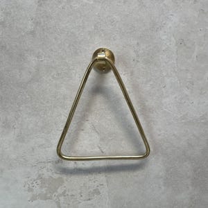 Op de afbeelding: Een goudkleurige, driehoekige handdoekring, gemonteerd op een cirkelvormige basis. De handdoekring is gemaakt van metaal en heeft een eenvoudig, modern ontwerp. De muur achter de ring is lichtgrijs.