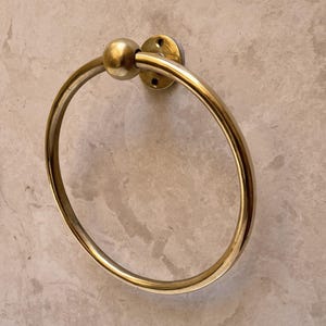 May include: A gold-colored towel ring with a circular design. The ring is attached to a round, flat base with screw holes. A small, spherical accent sits at the top of the ring. The hardware is mounted on a light gray, textured wall.