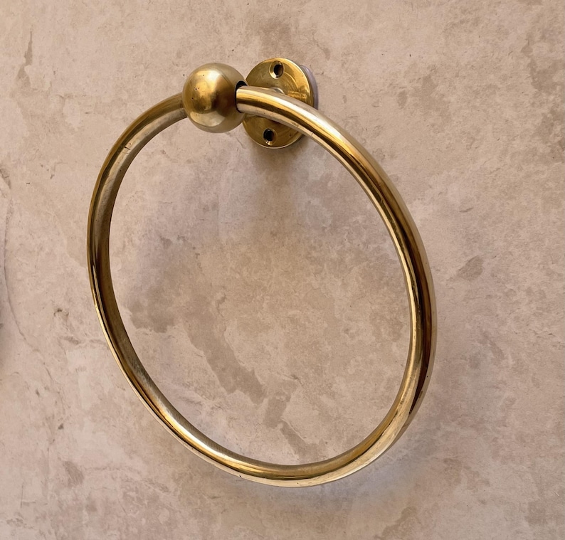 May include: A gold-colored towel ring with a circular design. The ring is attached to a round, flat base with screw holes. A small, spherical accent sits at the top of the ring. The hardware is mounted on a light gray, textured wall.