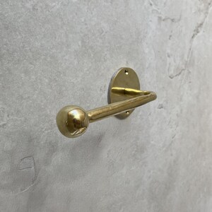 May include: A brass-colored wall hook with a rounded ball end. The hook is mounted on a circular plate with three screw holes. The hook is made of metal and is designed to hold items.
