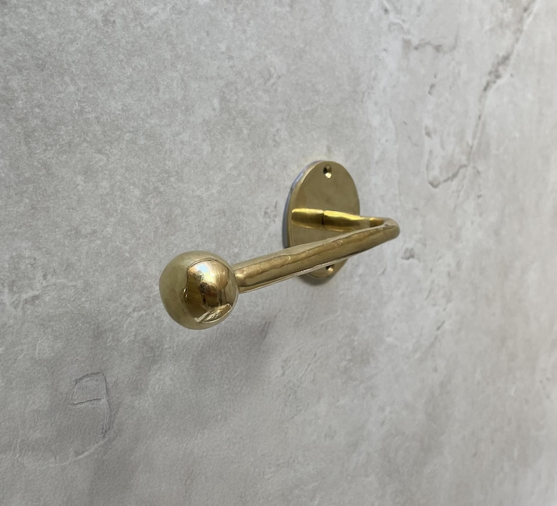 May include: A brass-colored wall hook with a rounded ball end. The hook is mounted on a circular plate with three screw holes. The hook is made of metal and is designed to hold items.