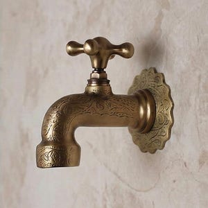 Pode incluir: Uma torneira de latão em estilo vintage com um design floral ornamentado. A torneira apresenta uma pega em forma de cruz e uma placa traseira decorativa. A torneira está montada numa parede de cor clara.