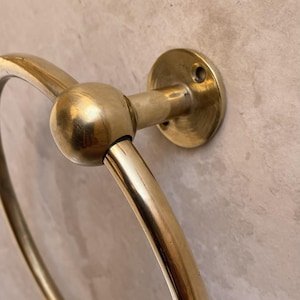 May include: A close-up of a brass towel ring. The ring is circular with a spherical joint connecting it to a circular wall mount. The brass has a polished, reflective finish, and the wall mount has three screw holes.