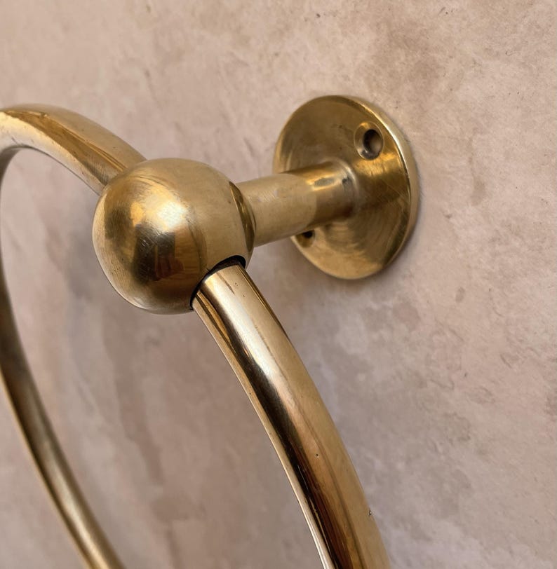 May include: A close-up of a brass towel ring. The ring is circular with a spherical joint connecting it to a circular wall mount. The brass has a polished, reflective finish, and the wall mount has three screw holes.