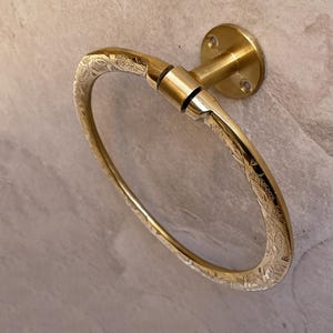 Pu&ograve; includere: Un portasciugamani dorato a forma ovale con motivo floreale. L'anello &egrave; montato su una base rotonda con fori per viti. Il metallo ha una finitura lucida e il disegno &egrave; inciso sulla superficie.