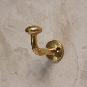 May include: A brass wall hook with a curved arm and a rounded knob. The hook is mounted on a circular base with a single screw hole. The finish is a warm, polished gold.