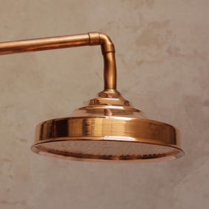 May include: A copper shower head with a round, ridged design. The shower head is attached to a copper pipe and elbow joint. The shower head has a textured surface and is made of copper.