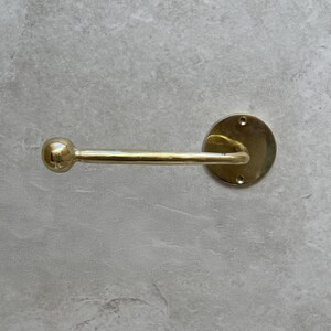 May include: A brass-colored towel bar with a round backplate and a straight bar ending in a small ball. The bar is mounted on a light gray, textured surface. The fixture has a classic, minimalist design.