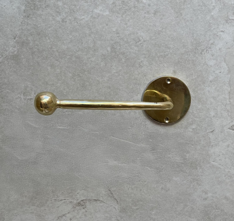 May include: A brass-colored towel bar with a round backplate and a straight bar ending in a small ball. The bar is mounted on a light gray, textured surface. The fixture has a classic, minimalist design.