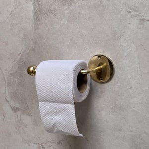May include: A brass toilet paper holder with a roll of white toilet paper. The holder is mounted on a light gray, textured wall. The holder has a circular base and a curved arm to hold the roll.
