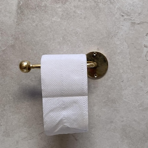 May include: A gold-toned toilet paper holder with a roll of white toilet paper. The holder has a round backplate and a curved bar to hold the paper. The wall is a light gray color.
