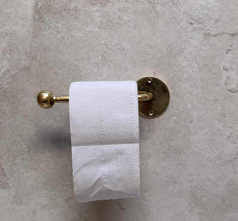 May include: A gold-toned toilet paper holder with a roll of white toilet paper. The holder has a round backplate and a curved bar to hold the paper. The wall is a light gray color.