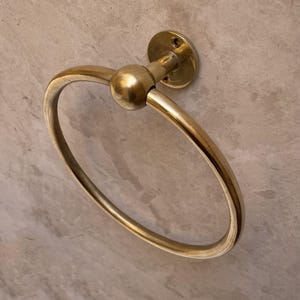 May include: A gold-colored, circular towel ring with a ball-shaped accent. The ring is attached to a round wall mount. The design is simple and elegant, suitable for a bathroom or powder room.