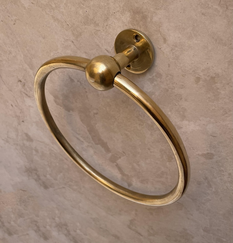 May include: A gold-colored, circular towel ring with a ball-shaped accent. The ring is attached to a round wall mount. The design is simple and elegant, suitable for a bathroom or powder room.
